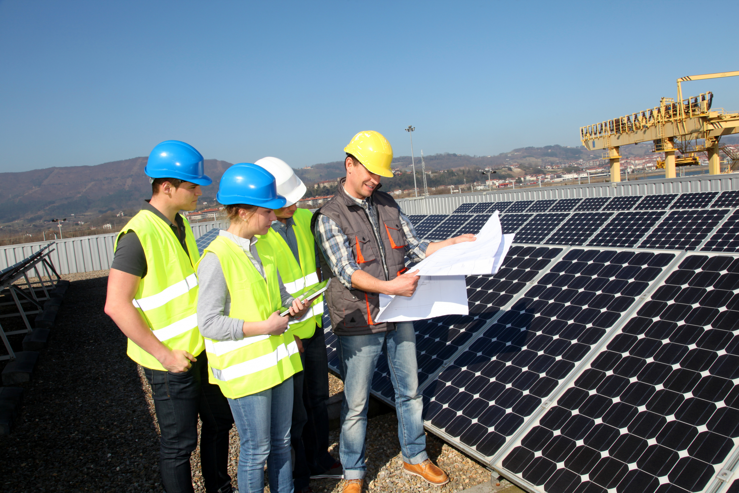 Group of people in yellow vest and hard hats standing and looking at a large piece of paper beside a solar panel.